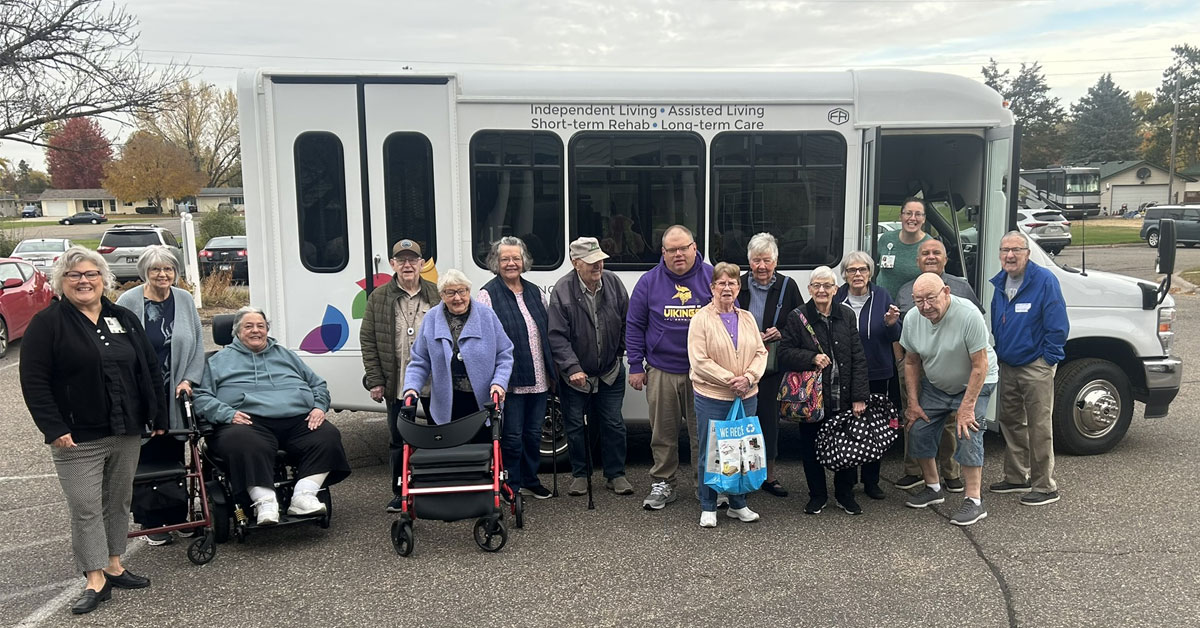 Group of senior living residents and staff standing together outdoors in front of a bus labeled for independent living, assisted living, and rehab services, with some using walkers and holding bags.