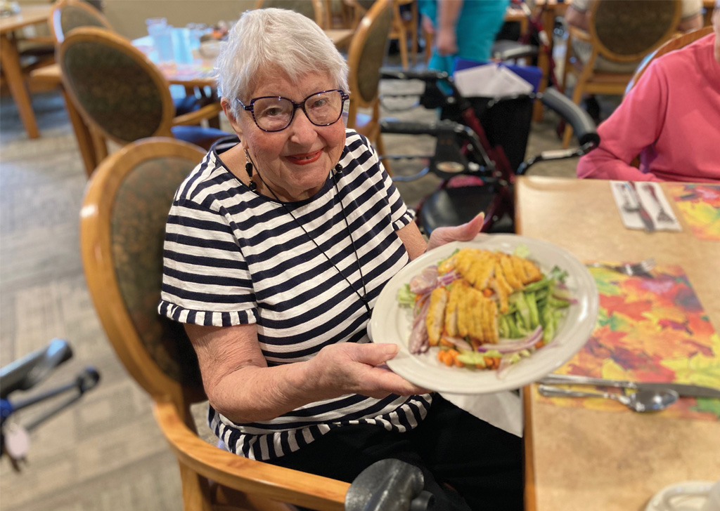 Resident seated at a dining table in a senior living community, holding a plate of grilled chicken salad with vegetables, with dining chairs and other residents visible in the background.