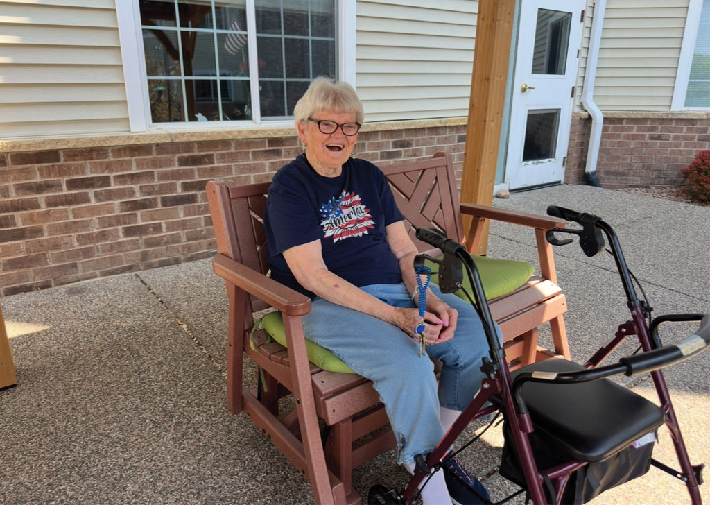 Resident seated on an outdoor bench at a senior living community, holding a walker nearby, with a brick building, large windows, and entry door in the background.